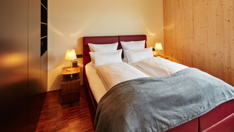 Hálószoba a Guntrams kerti padláson, © Herbert Lehmann Hangulatos hálószoba franciaággyal, piros fejvéggel, két éjjeliszekrénnyel, lámpákkal és fából készült falakkal.