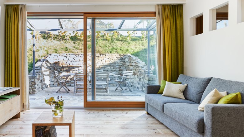 Weinviertler Kellerstöckl, © Andreas Hofer A modern nappali belső nézete, szürke kanapéval, fapadlóval és nagy ablakokkal, amelyek egy kőfallal és kerti bútorokkal berendezett teraszra vezetnek.