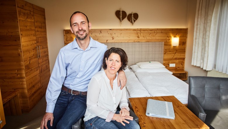 Szobák a Dissauer család Rosinger farmján, © Familie Rosinger Egy pár ül egy hangulatos, fából készült bútorokkal és franciaággyal berendezett szobában.