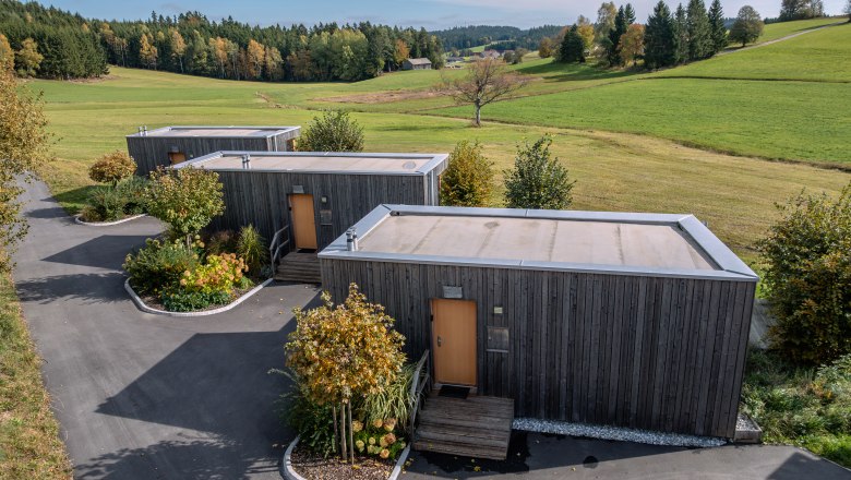 roo'n, © Niederösterreich Werbung / Maximilian Pawlikowsky Három modern faház egy zöld, fákkal és rétekkel tarkított tájban.