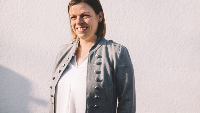 Szolgálatban: Sandra Buchinger-Löfler, © Niederösterreich Werbung/Mara Hohla Szürke kabátos nő egy fehér fal előtt.