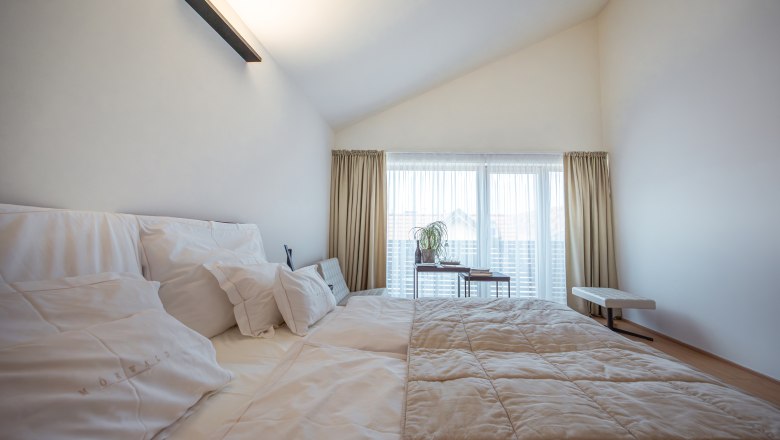 Relais & Chateaux MÖRWALD Hotel am Wagram, © Niederösterreich Werbung / Maximilian Pawlikowsky Modern szállodai szoba nagy ággyal, fehér párnákkal és ágytakaróval, világos függönyökkel és ablakkal a háttérben.