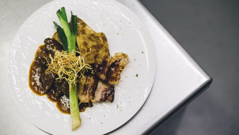 Marhasült az Alpenvorland Beiriedből, © Niederösterreich Werbung/David Schreiber Egy tányér sült marhahússal, burgonyagratinnal, póréhagymával és ropogós hagymával fém felületen.