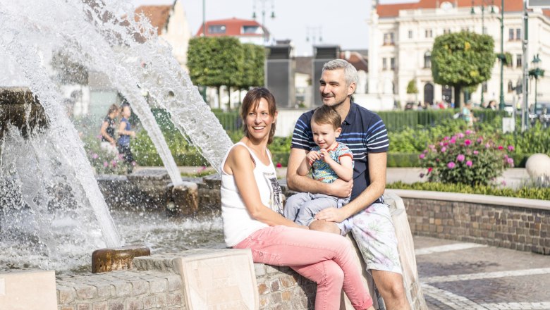 c-stadtgemeinde-tulln-robert-herbst-20, © Hauptplatz-Tulln-c-Stadtgemeinde Robert-Herbst-20 Eine Familie sitzt an einem Brunnen in einer Stadt mit historischen Gebäuden im Hintergrund.