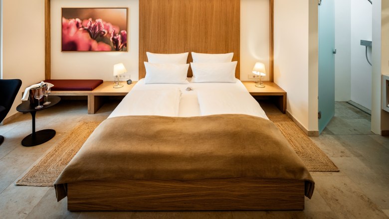 Vendégház Flora, © Herbert Lehmann Modernül berendezett szállodai szoba franciaággyal, éjjeliszekrényekkel, lámpákkal és egy képpel a falon.