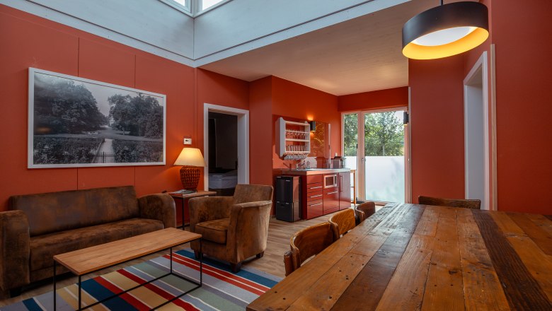 Grafenegg nyaralók, © Niederösterreich Werbung / Maximilian Pawlikowsky Belső nézet egy modern nappaliból, vörös falakkal, fa asztallal, kanapéval és konyhasarokkal.