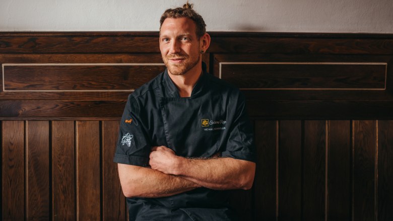 Michael Schindler bérbeadó, © Niederösterreich Werbung/Daniela Führer Egy fekete szakácskabátos férfi ül egy fából készült fal előtt.