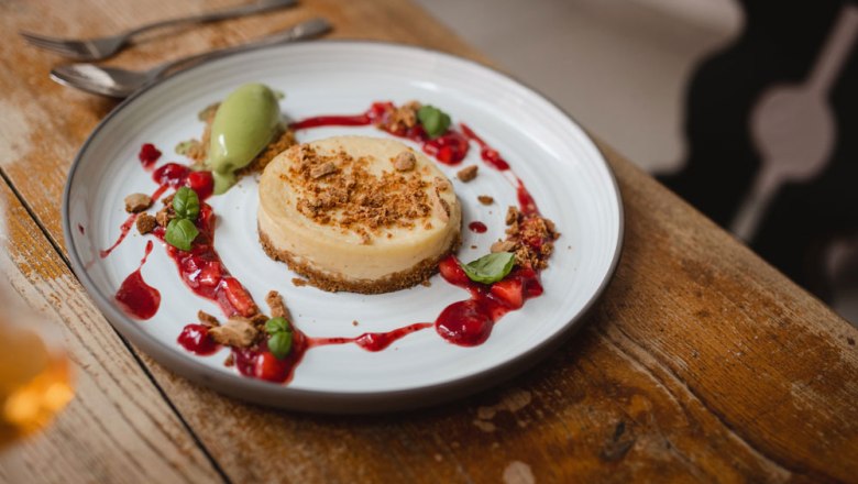 Sajtos sütemény, © Niederösterreich Werbung/Daniela Führer Egy tányéron piros bogyókkal és zöld fagylalttal díszített sajttorta egy faasztalon.