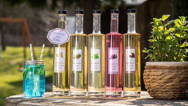 Házi szörpök a Rath-Smetana fogadó gyógynövénykertjéből, © Niederösterreich Werbung/Kurt Pinter Öt üveg házi készítésű szörp és egy kék pohár szívószállal egy kinti faasztalon.