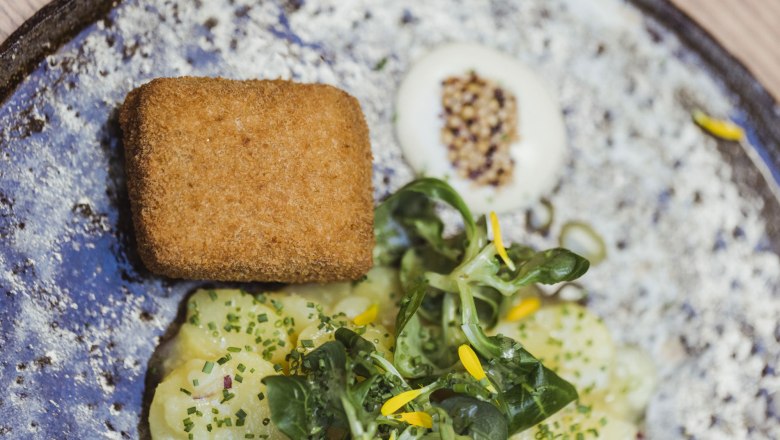 Borjúfej madárfejen és almasalátán, © Niederösterreich Werbung/David Schreiber Egy tányér rántott borjúfej, burgonyasaláta és báránysaláta, sárga virágokkal díszítve.