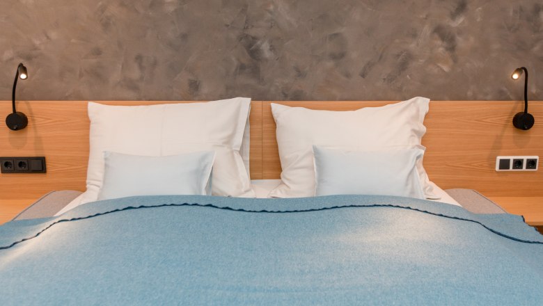 Ház a tónál, © schwarz-koenig.at Egy franciaágy, kék takaróval és fehér párnákkal egy szürke fal előtt.