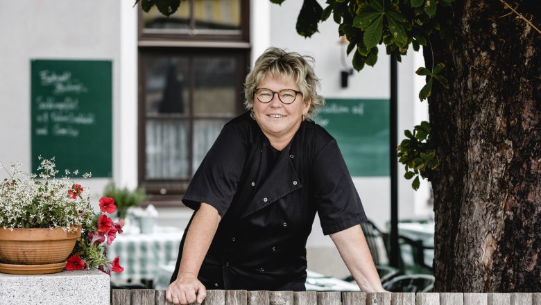 Martina Ebner háziasszony, © Niederösterreich Werbung/David Schreiber Egy fekete ruhás nő egy fakerítésnek támaszkodik, a háttérben virágok és egy fa.