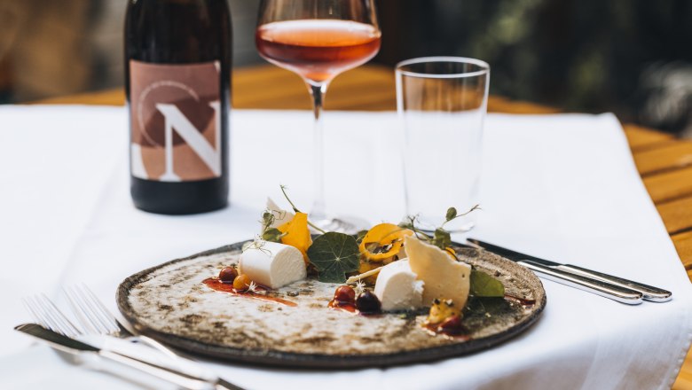 Saját borunk, © Niederösterreich Werbung/David Schreiber Egy tányér sajt és köret, egy pohár rozé bor és egy üveg bor az asztalon.