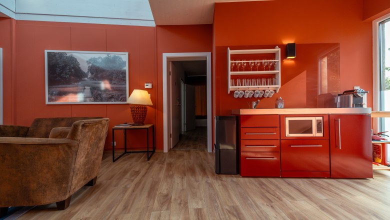 Grafenegg nyaralók, © Niederösterreich Werbung / Maximilian Pawlikowsky Belső nézet egy modern nappaliból, piros falakkal, piros konyhabútorral, barna fotelekkel és egy nagy ablakkal.