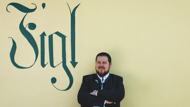 Matthias Strunz, a Figl bérbeadója, © Niederösterreich Werbung/David Schreiber Egy hagyományos öltözetű férfi áll egy sárga fal előtt, amelyre a "Figl" szó van írva.