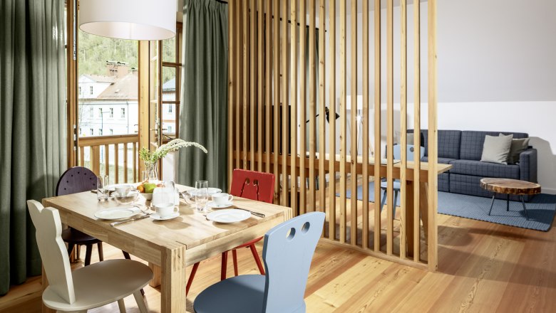 Hotel zum Glockenturm, © www.croce.at Világos szállodai szoba, fapadlóval, étkezőasztallal és székekkel, amelyet egy fa lécfal választ el, egy kanapéra és egy dohányzóasztalra nyílik kilátás.