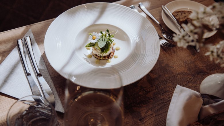 Füstölt pisztrángtatár burgonyás palacsintán, © Niederösterreich Werbung/thecreatingclick.com Egy tányér füstölt pisztrángtatár burgonyapalacsintán, fűszernövényekkel és virágokkal díszítve, egy faasztalon evőeszközökkel és egy pohár borral.
