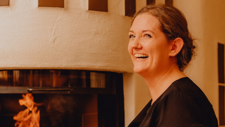 Eva Maria Reisenbauer házinéni, © Niederösterreich Werbung/Klaus Engelmayer Nő mosolyog egy kandalló előtt, ahol ég a tűz.