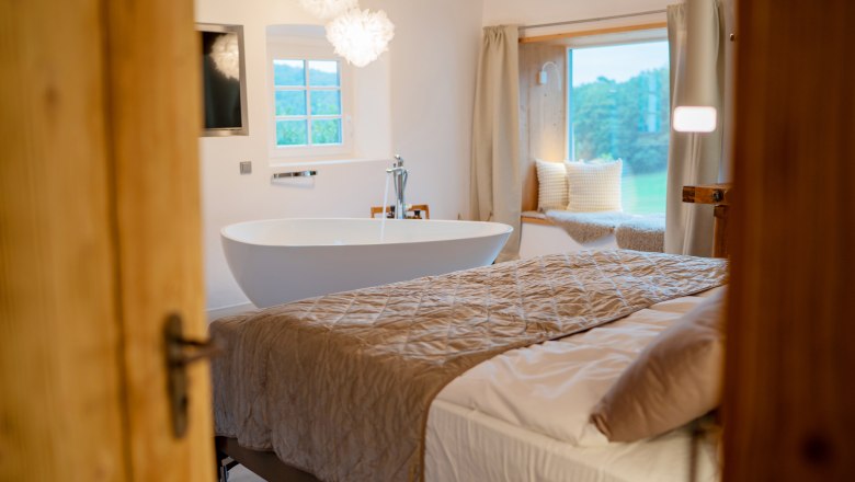 Csodákkal teli faházak és helyek - Weidehaus De Luxe, © WUNDERVOLL eben Belső nézet egy luxus hálószobából, szabadon álló káddal, nagy ággyal és párnákkal ellátott ablakpárnával.