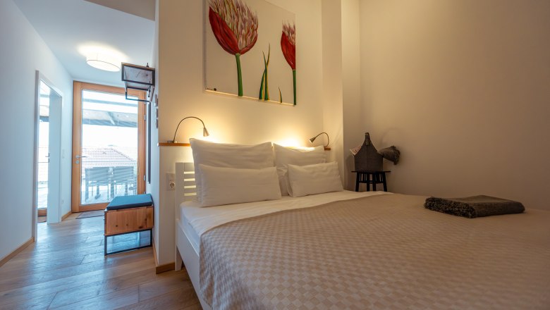 Hausgnost Apartmanok, © Niederösterreich Werbung / Maximilian Pawlikowsky Modern hálószoba franciaággyal, világos fapadlóval és művészeti alkotásokkal a falon. Az ajtó a háztetőkre néző erkélyre vezet.