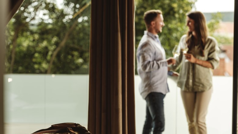 Pincészet Hagn Kellerstöckl, © Weinviertel Tourismus GmbH Egy pár áll egy erkélyen, kezében pohár borral, az előtérben egy utazótáska fekszik egy ágyon.