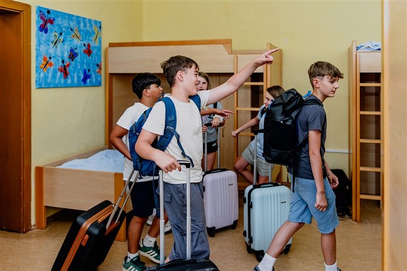 Gyerekcsoport bőröndökkel egy emeletes ágyakkal felszerelt hálóteremben.