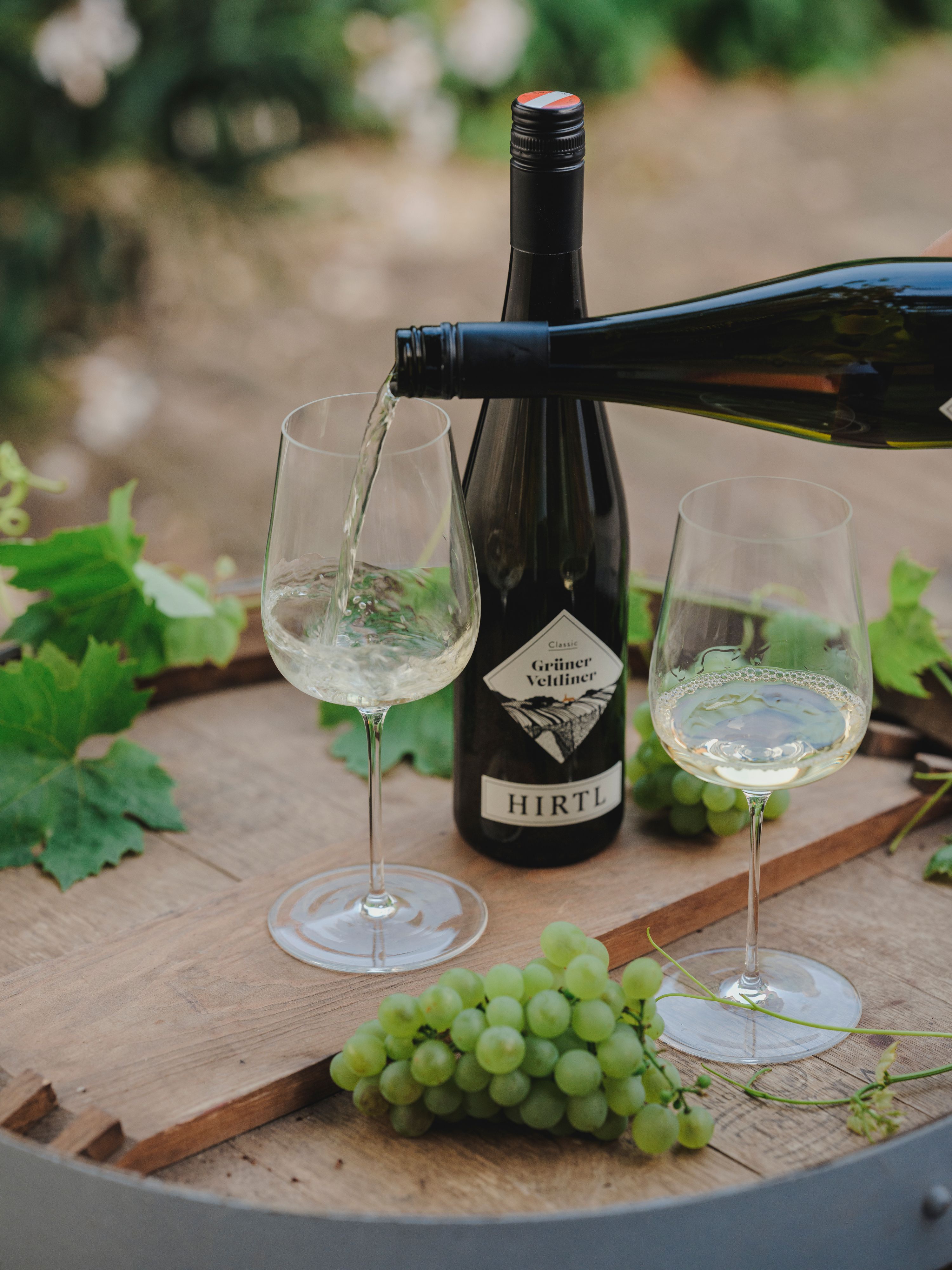 Fehérbort töltenek egy pohárba, mellette egy üveg Zöldveltelini Hirtlből és zöld szőlő egy faasztalon.