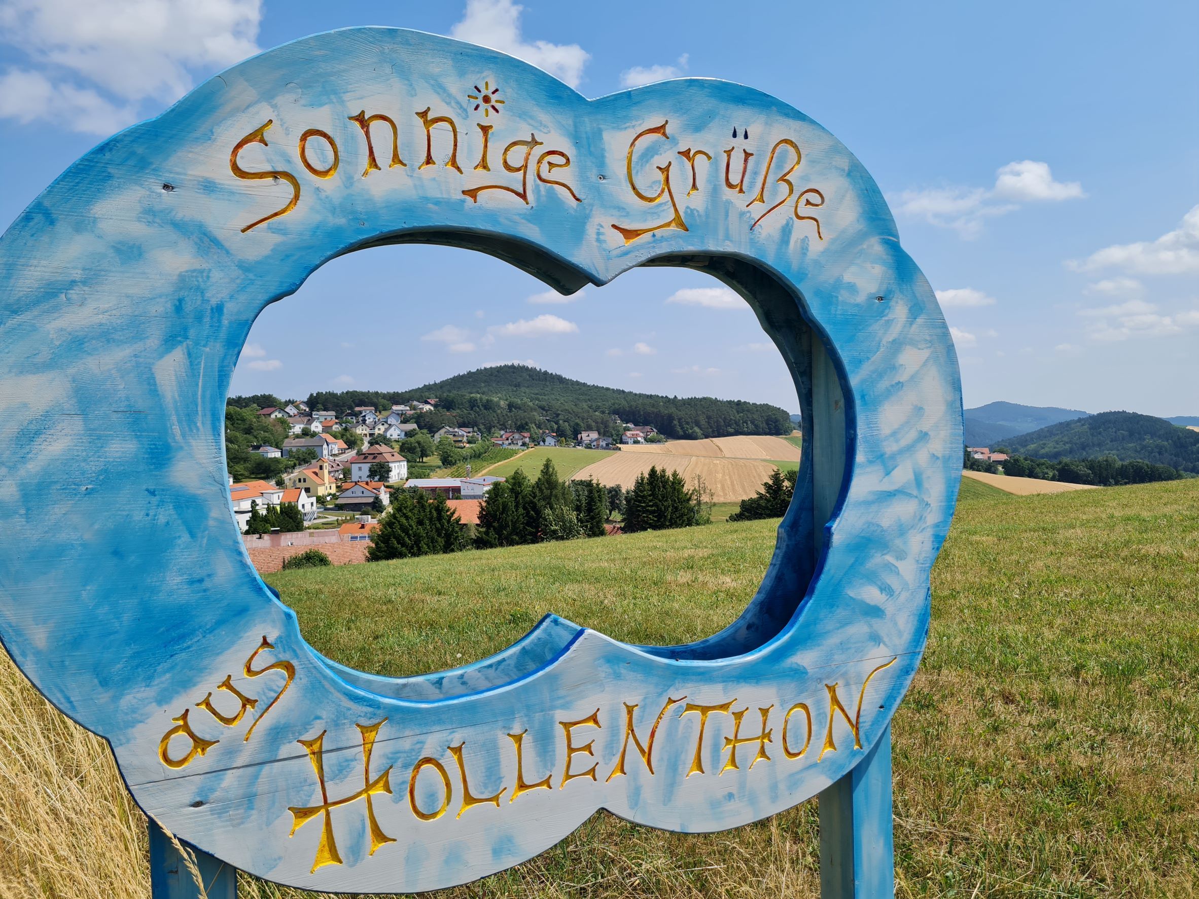 Kék fakeret felhő alakú felirattal: "Sunny greetings from Hollenthon" (Napos üdvözlet Hollenthonból) egy tájkép előtt.