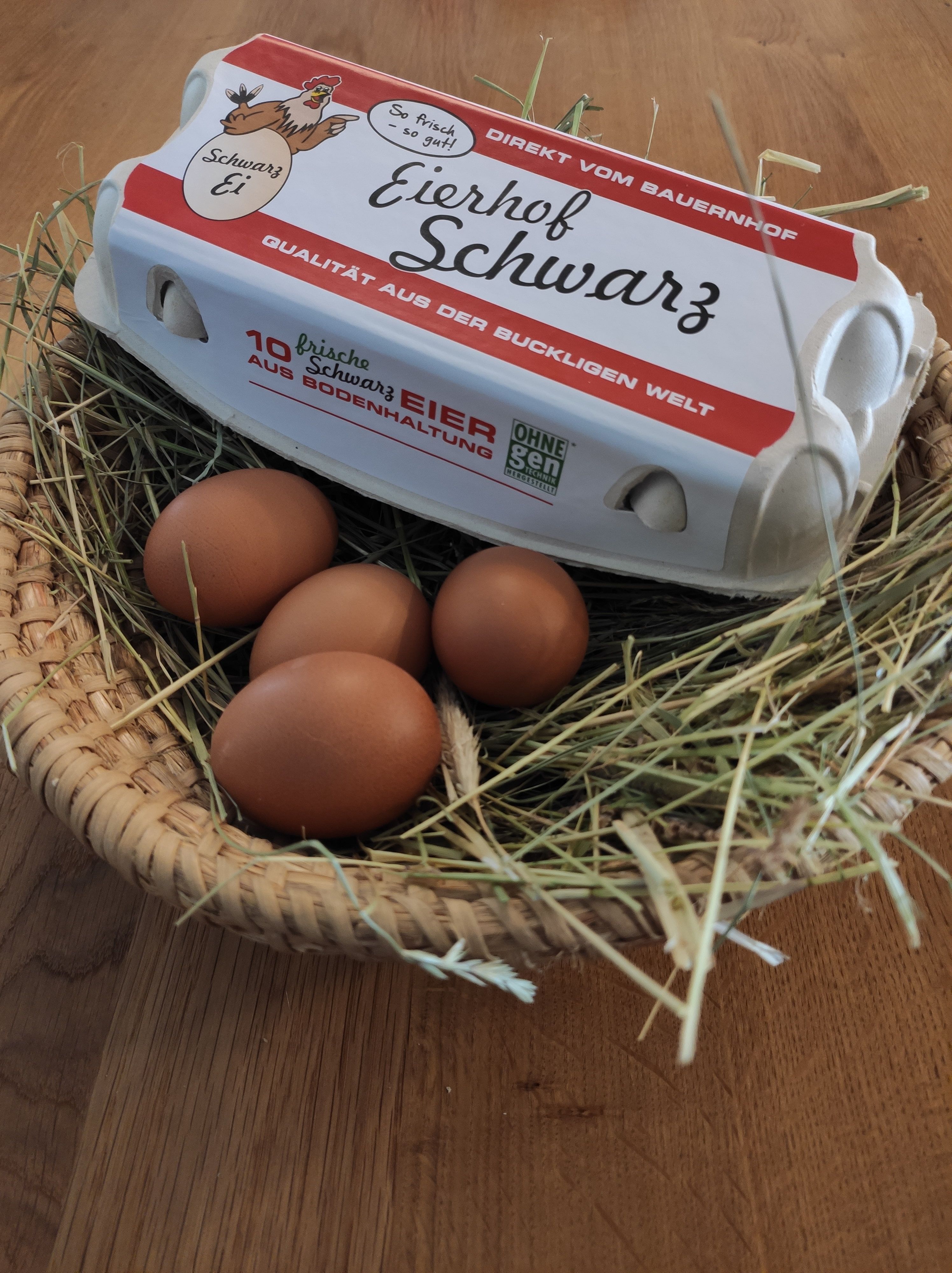 Egy "Eierhof Schwarz" feliratú tojástartó doboz fekszik egy szalmával és barna tojásokkal teli kosáron.