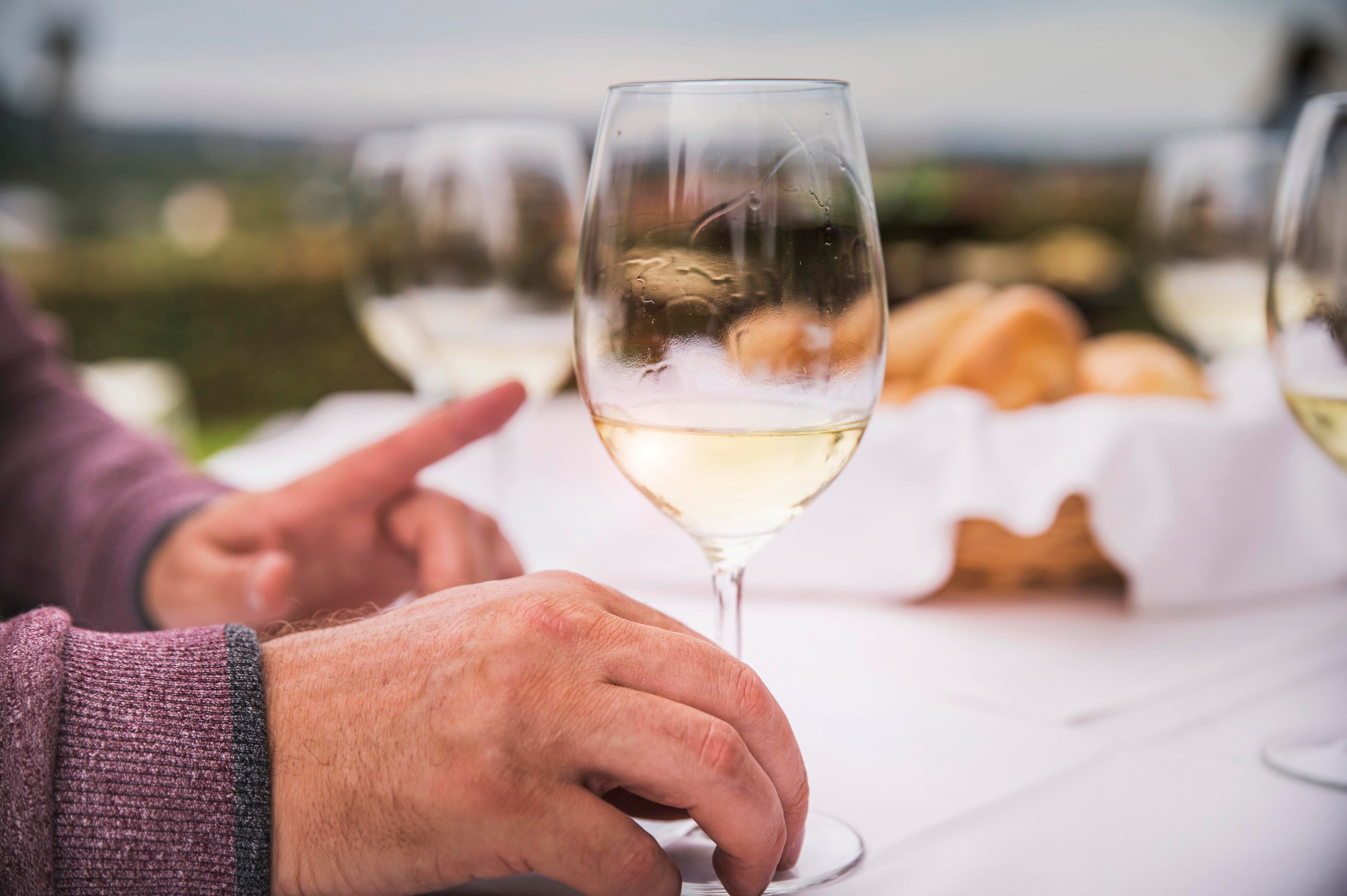 Közelkép a kezekről borospohárral egy megterített asztalon a szabadban.