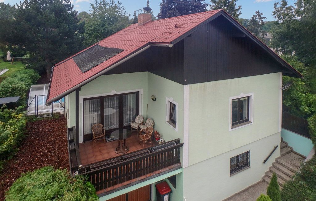 Kétemeletes ház piros tetővel és erkéllyel, fákkal körülvéve.