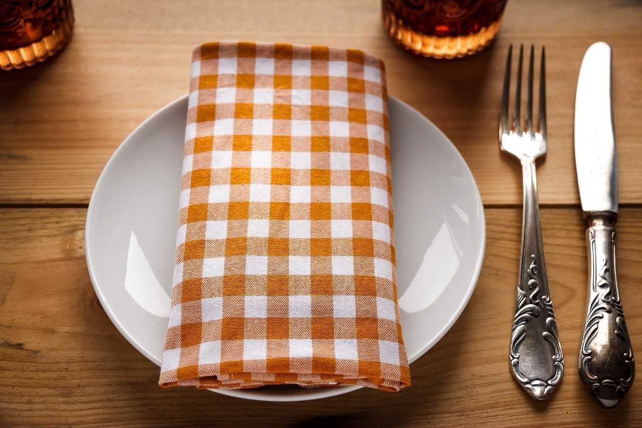Egy tányér narancs-fehér kockás szalvétával, mellette evőeszközök egy faasztalon.
