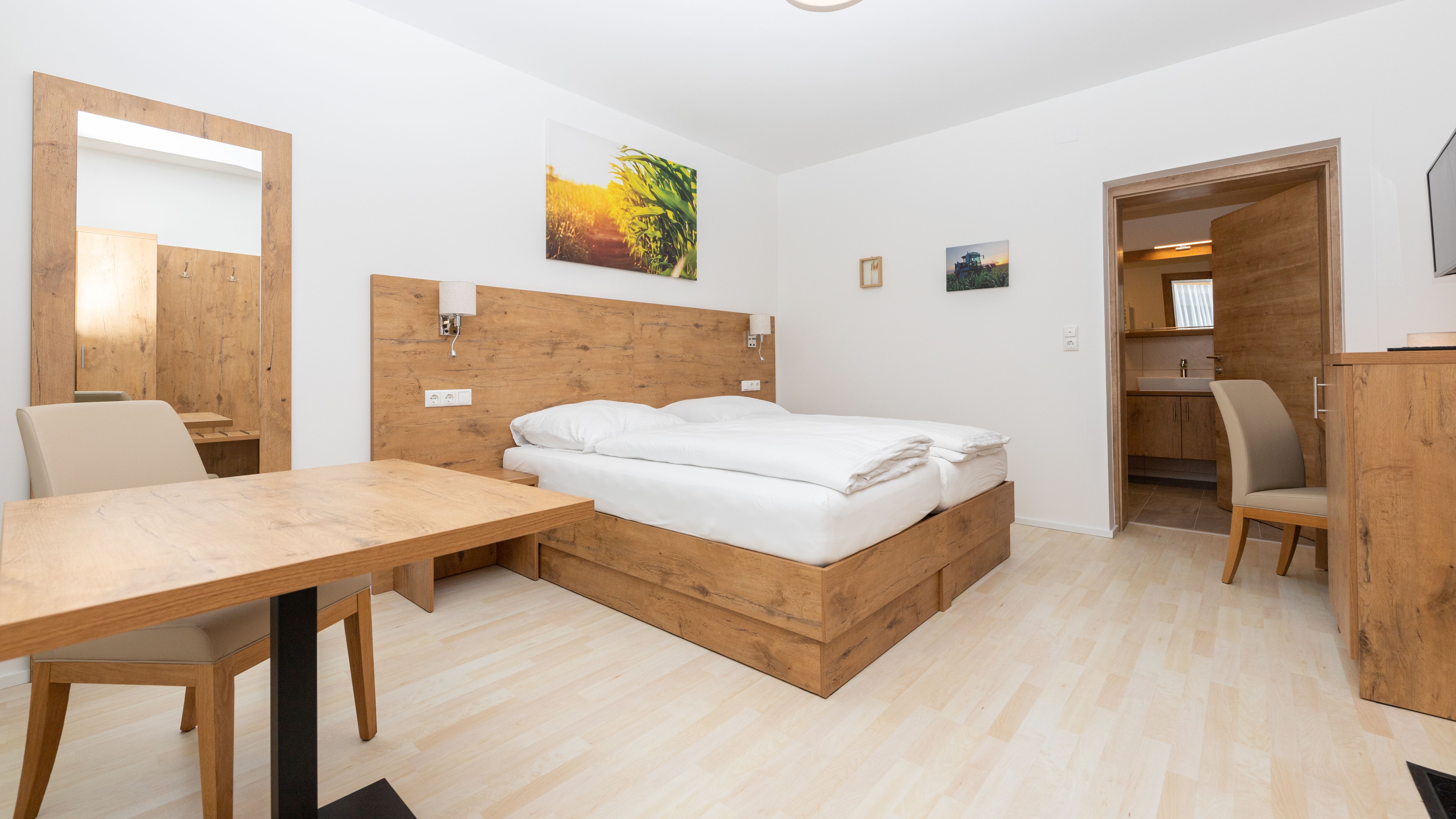 Modern hálószoba fabútorokkal, franciaággyal, asztallal és székkel. A falon tükör és képek, a fürdőszobába vezető ajtó.