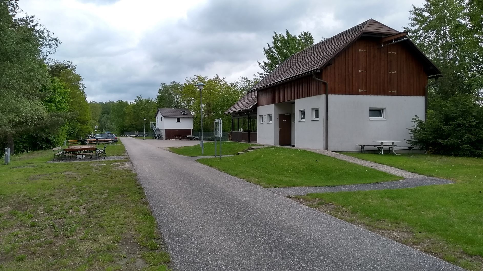 Egy ösvény vezet el egy fehér, faburkolatos épület mellett, amelyet fák és rétek vesznek körül, felhős égbolt alatt.