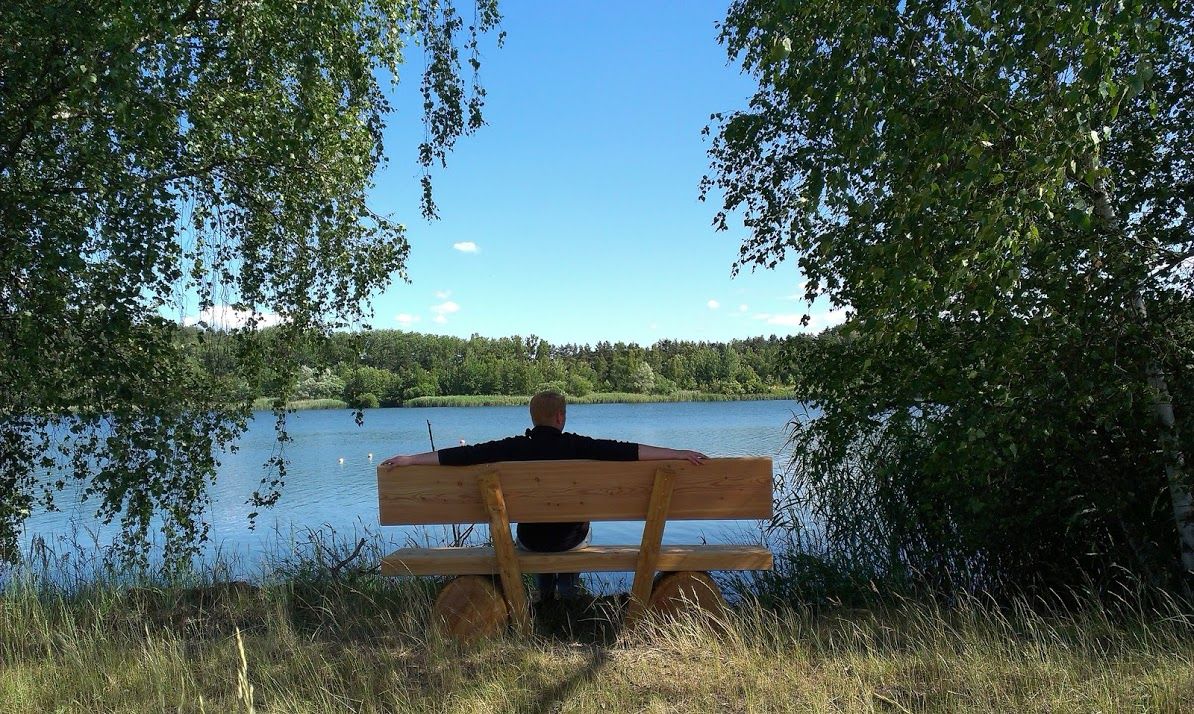 Egy tóparton, fákkal körülvett padon ülő személy.