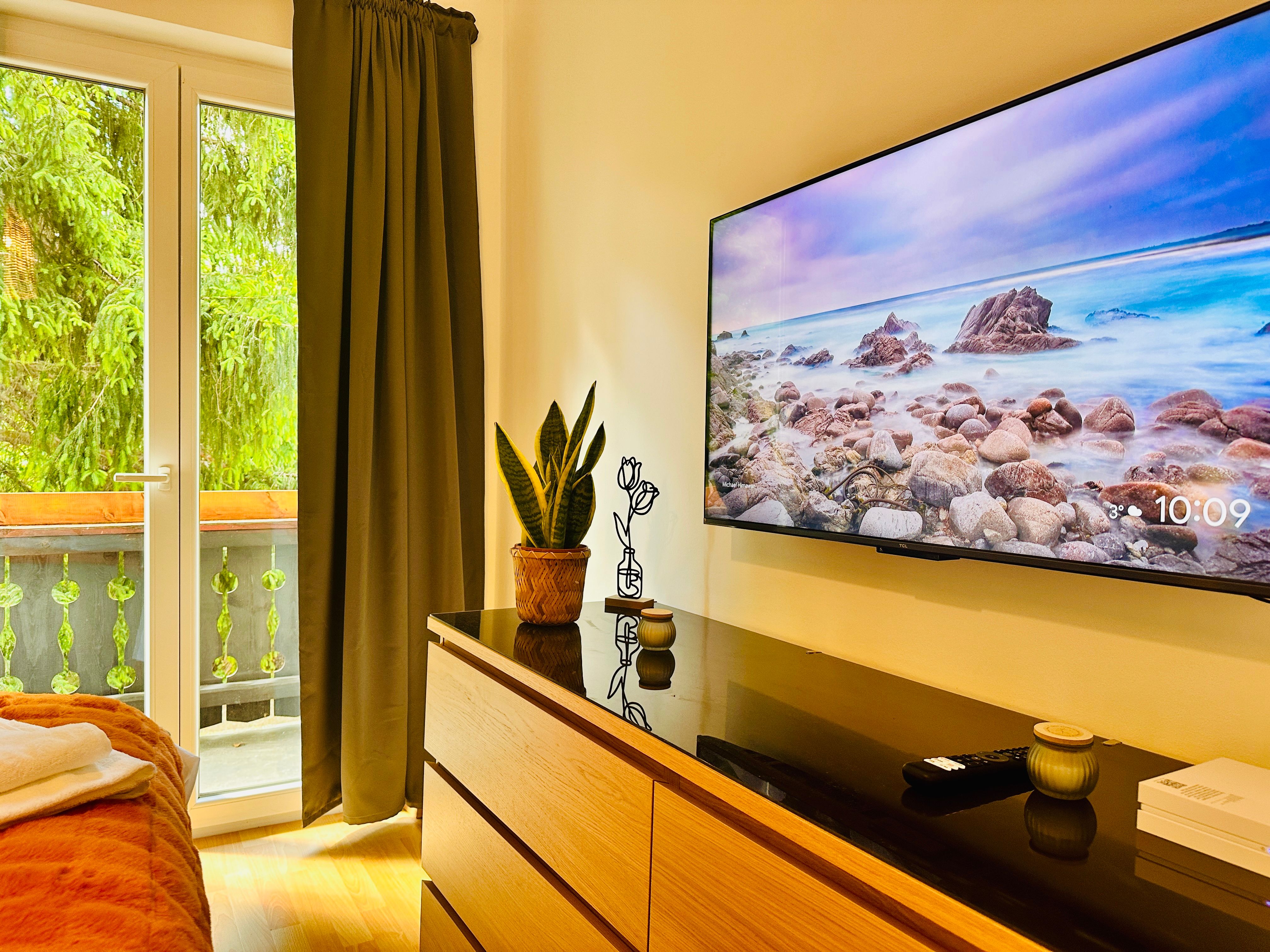 Hangulatos szoba TV-vel, fiókos szekrénnyel, növényekkel és erkélyre néző kilátással.