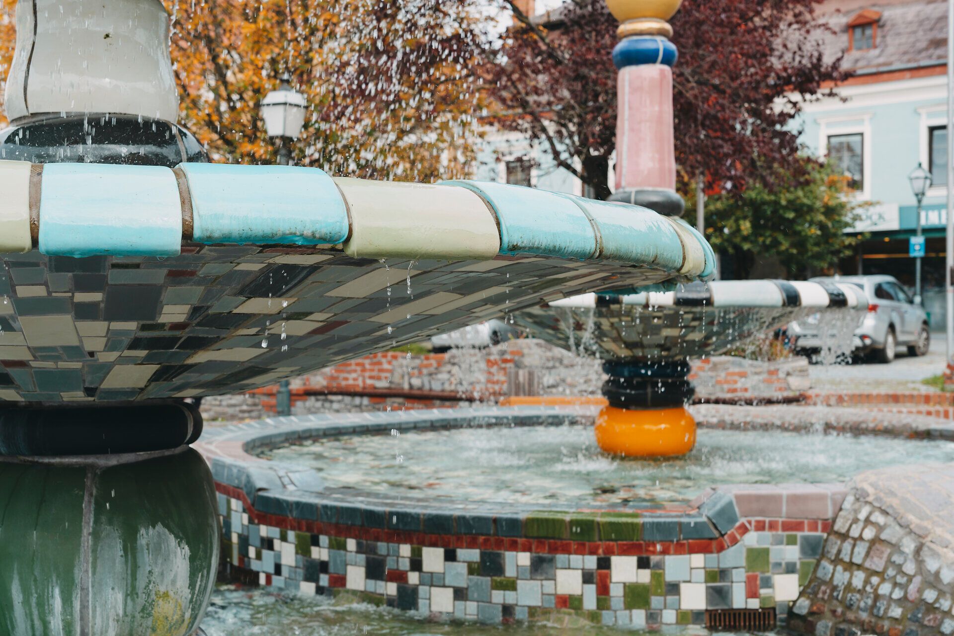 Az ősz különleges varázslatot kölcsönöz a Hundertwasser-kútnak, amikor a víz lágyan fodrozódik a színes csempéken. Az élénk színű fákkal és hívogató hangulattal körülvett hely elidőzésre és álmodozásra invitál.