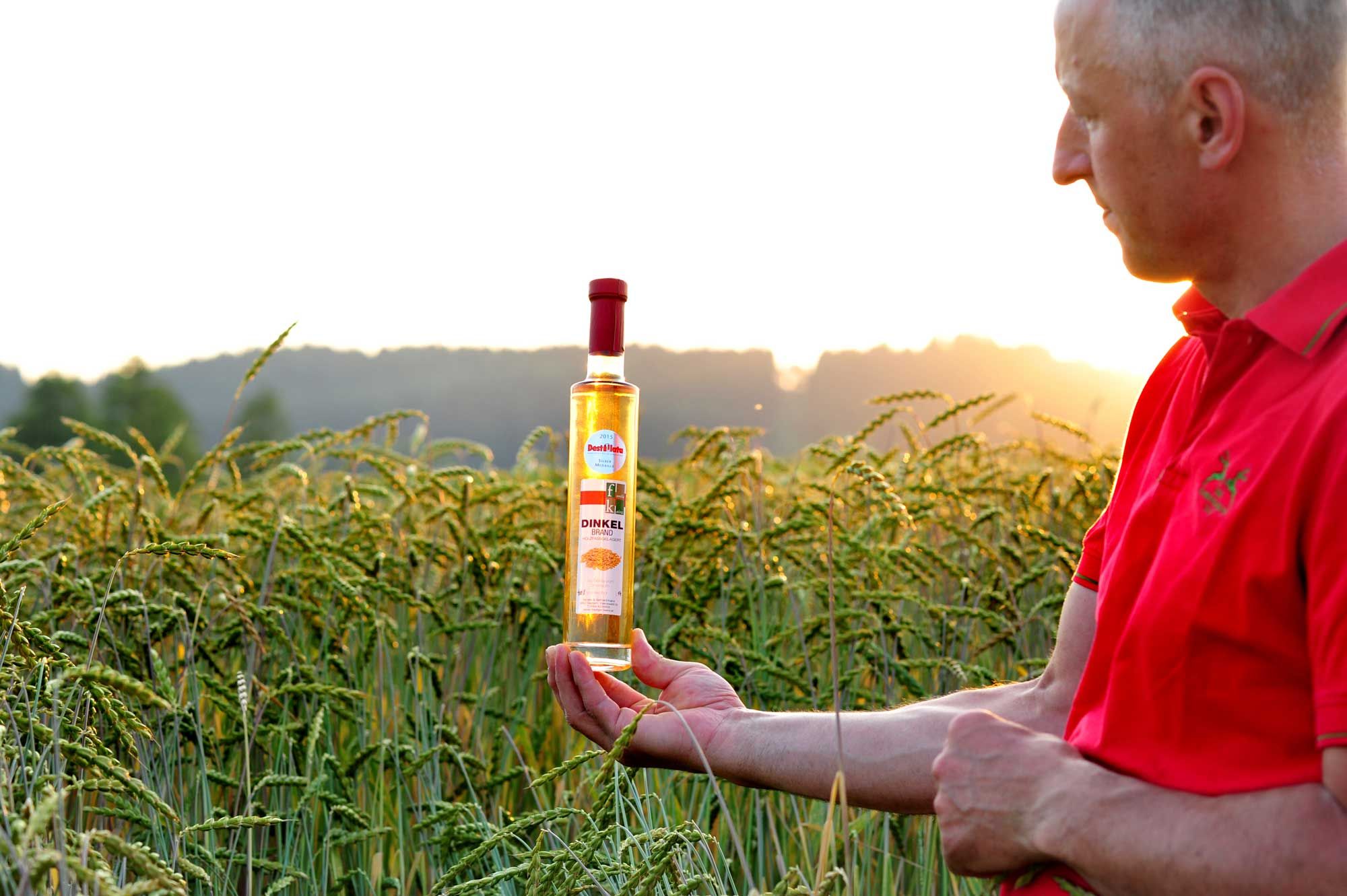 Egy piros inges férfi egy üveg tönkölypálinkát tart a kezében egy kukoricamezőn naplementekor.
