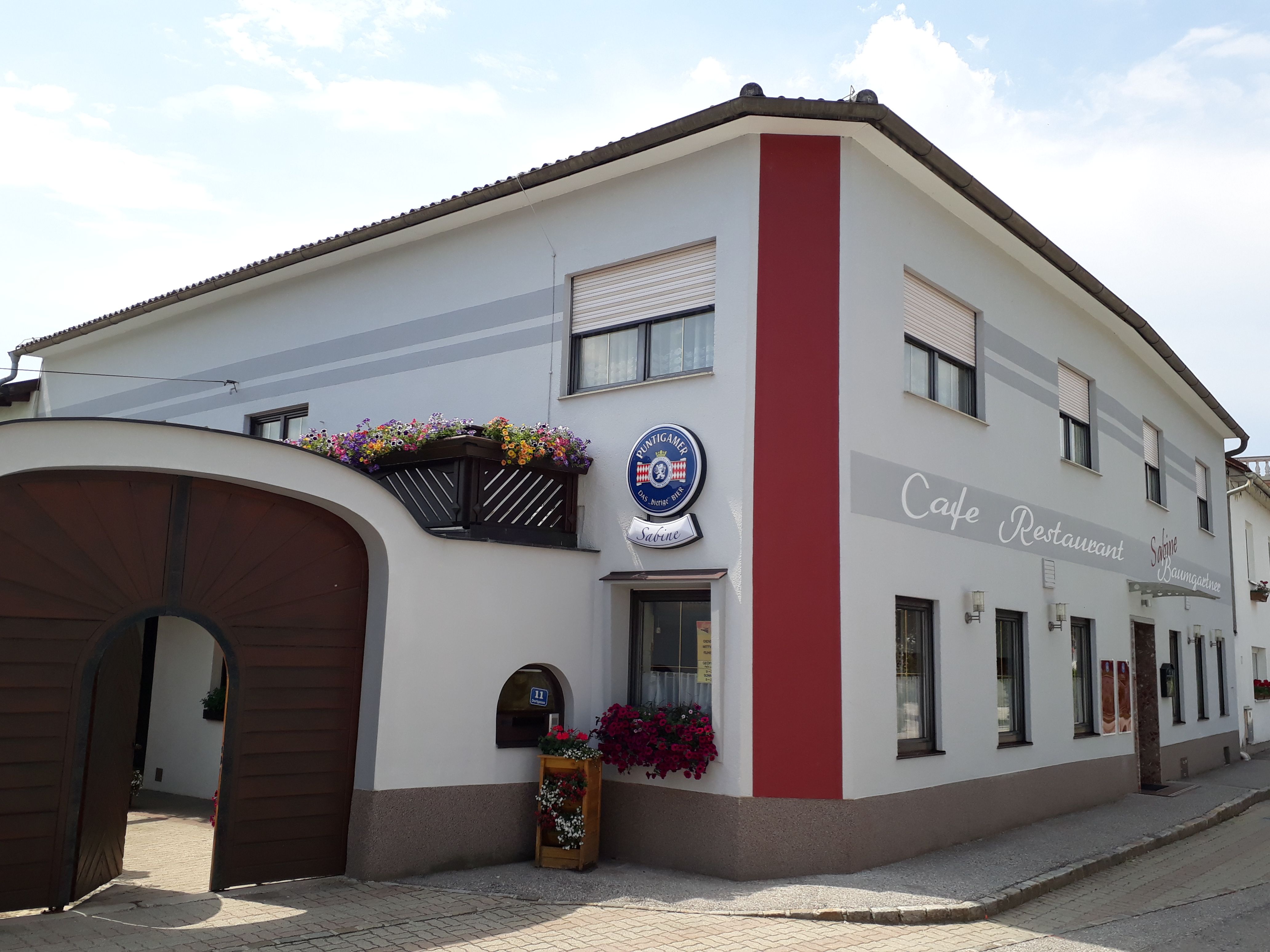 A Café Sabine külső nézete fakapuval, fehér és piros homlokzattal és a "Café-Restaurant" felirattal.