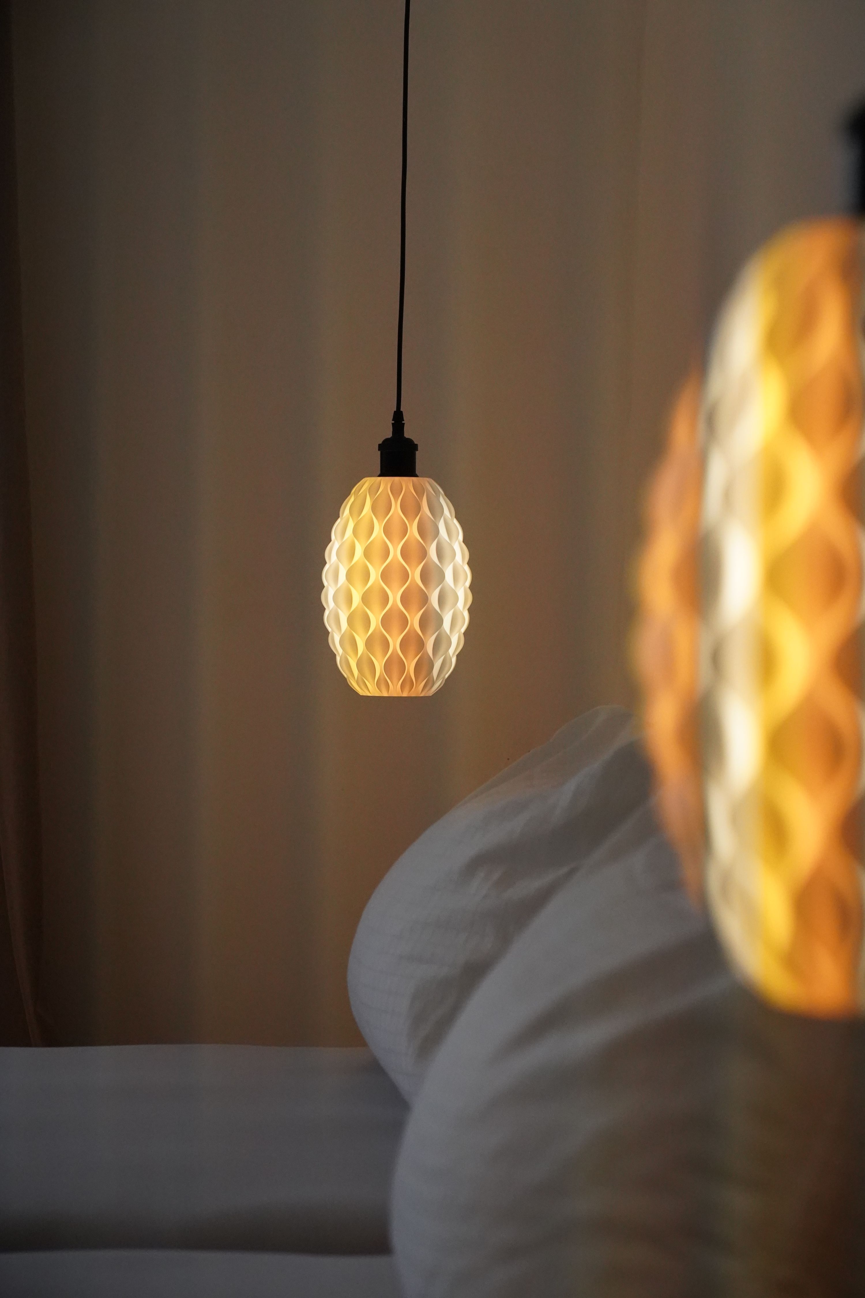 Közelkép egy meleg fényt sugárzó, méhsejtmintás függőlámpáról egy ágy mellett.