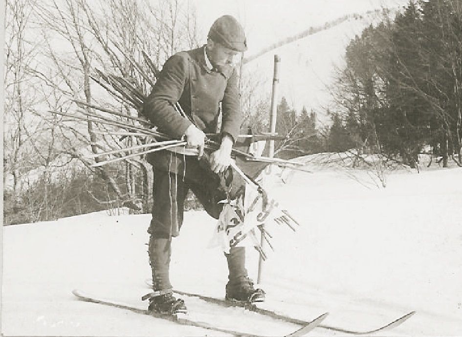 Történelmi fénykép egy síelő férfiról, síbotokkal a hóban.