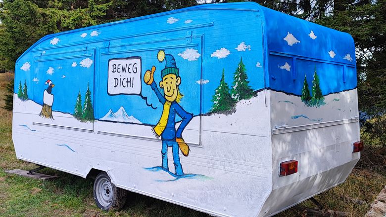 Színesen festett lakókocsi, téli tájjal és egy "Mozgás!" feliratú figurával.