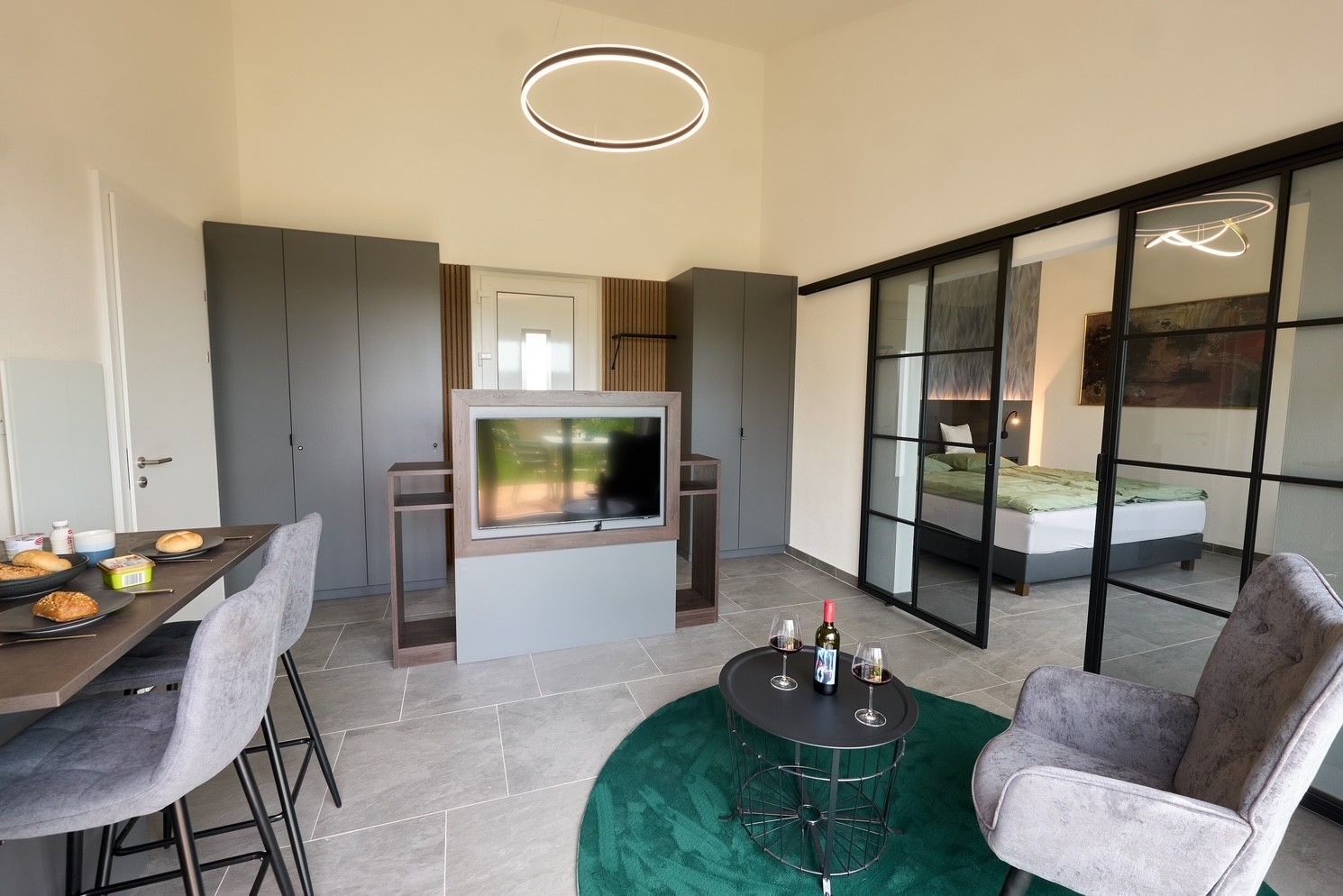 Modern lakás étkezőasztallal, TV-vel, szürke szekrényekkel és üveg tolóajtókkal a hálószobába.