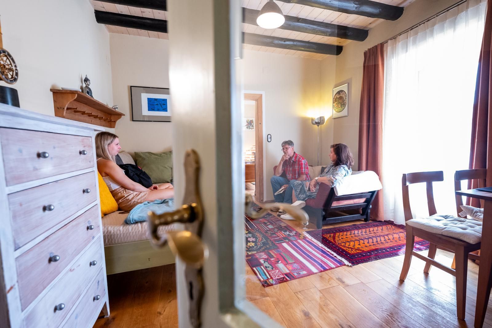 Három ember ül egy hangulatos, fapadlóval és színes szőnyeggel borított nappaliban.