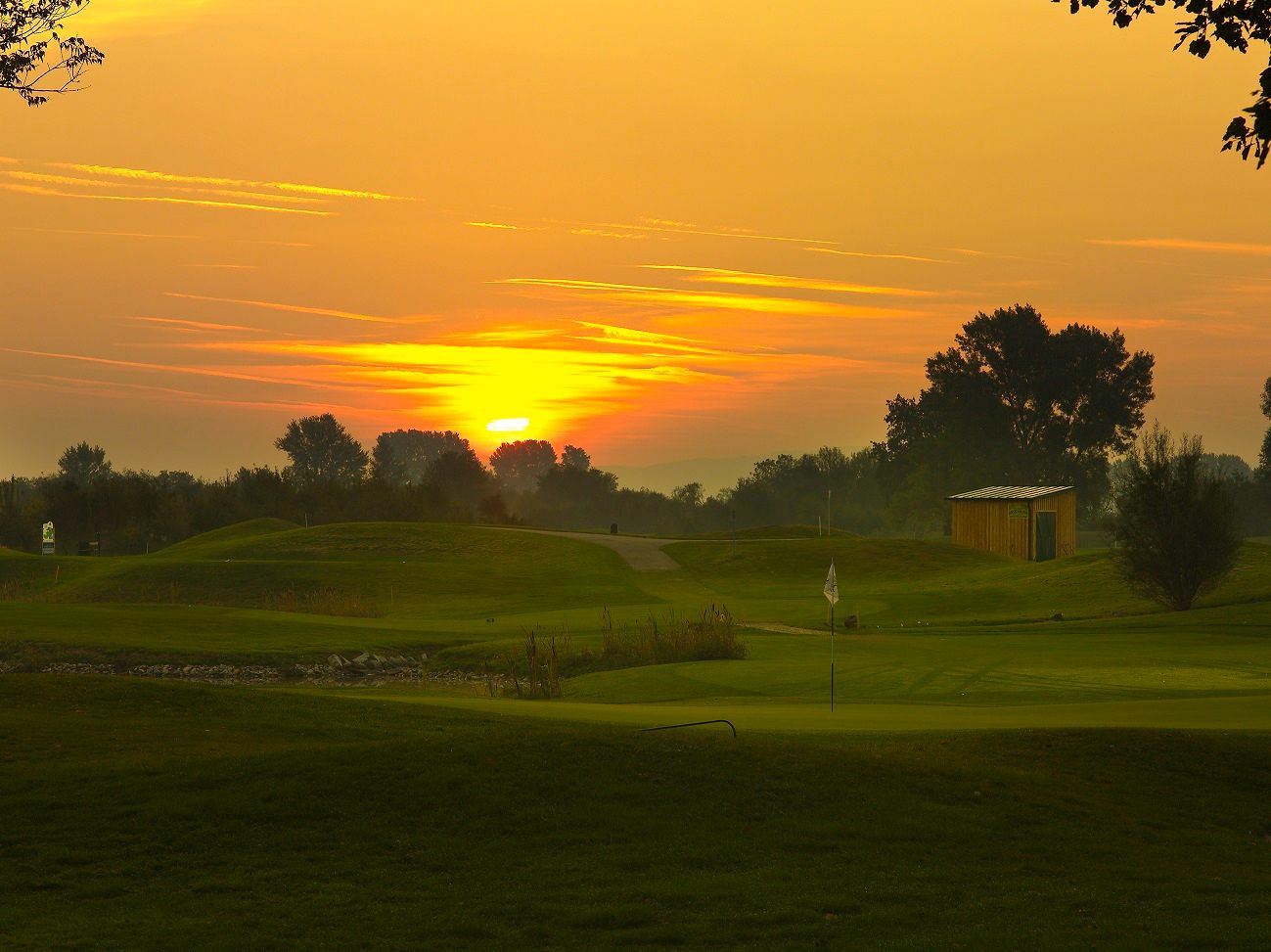Naplemente egy golfpálya felett, zászlóval és kis épülettel.