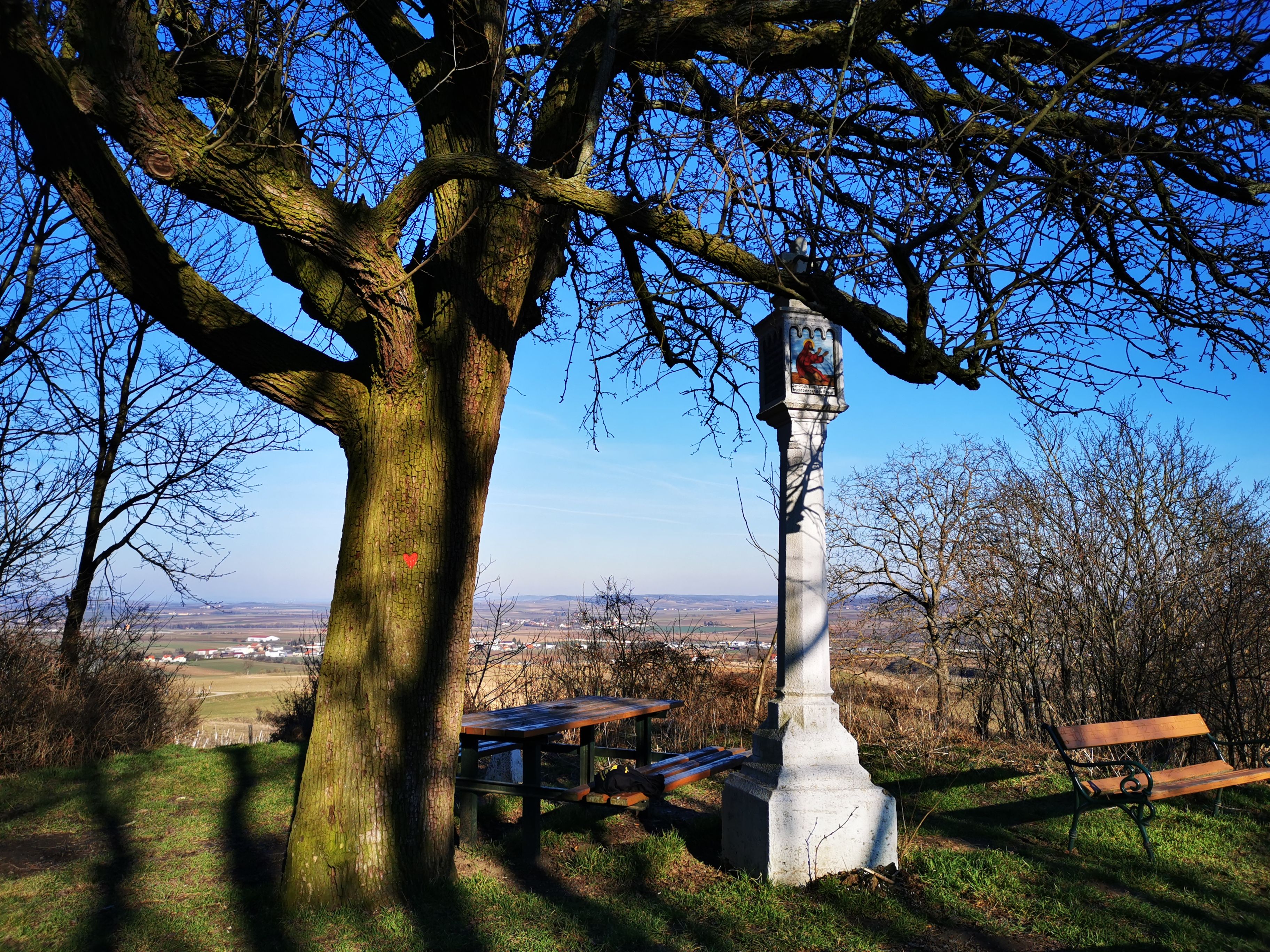Egy nagy fa mellett egy kő útmenti kereszt, padokkal és a tájra nyíló panorámával.