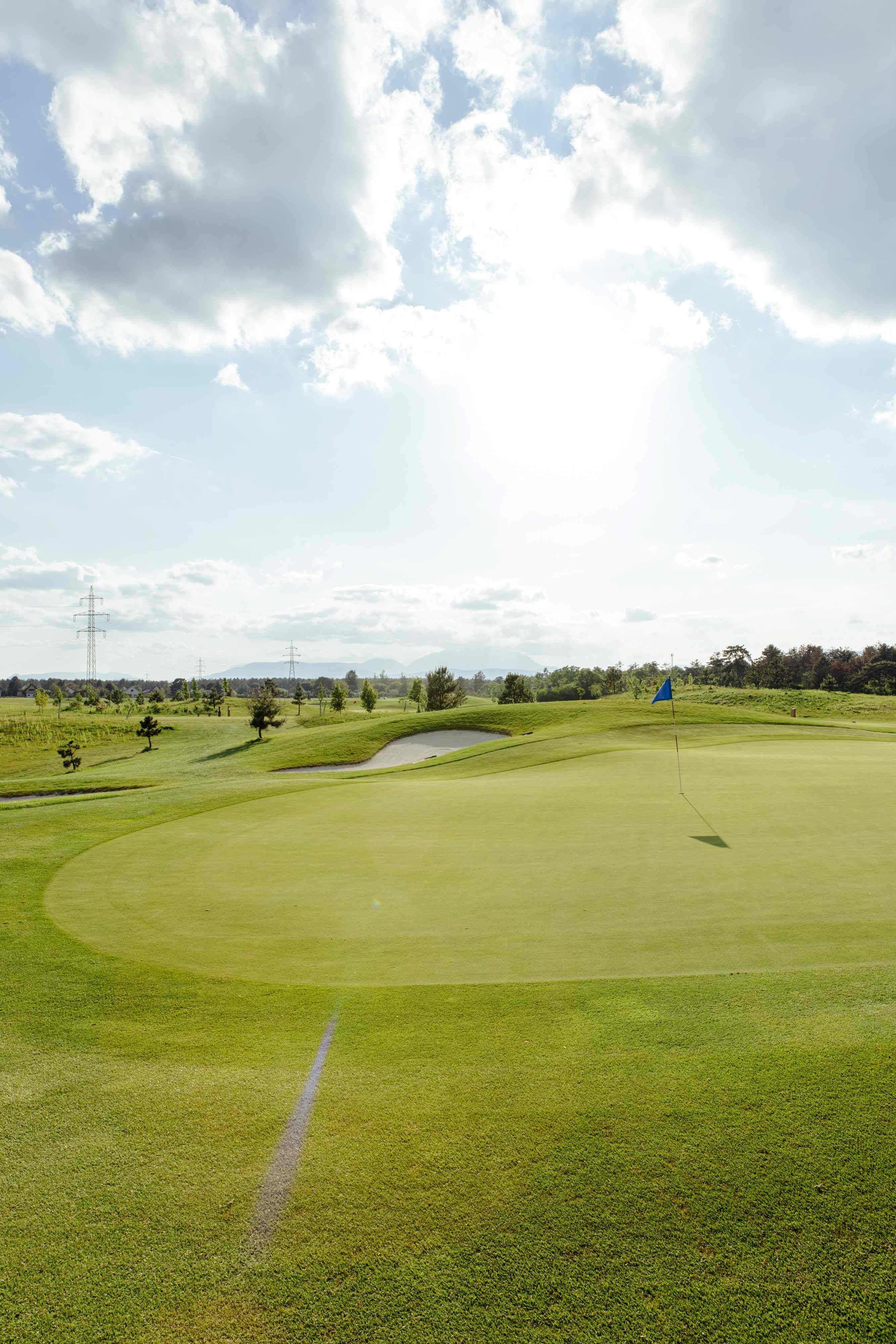 Golfpálya zászlóval a zöldön, fákkal és villanyoszlopokkal körülvéve, felhős égbolt alatt.