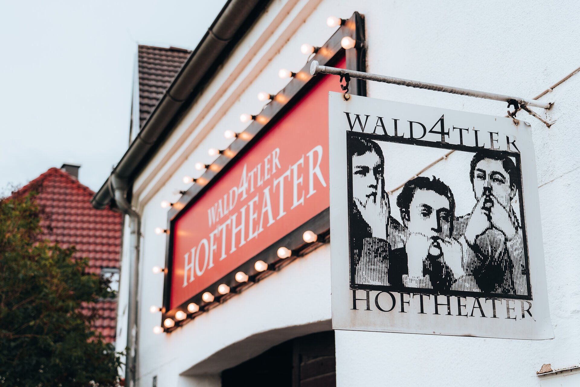 A Wald4tler Hoftheater külső nézete, világító táblával és három ember képével.