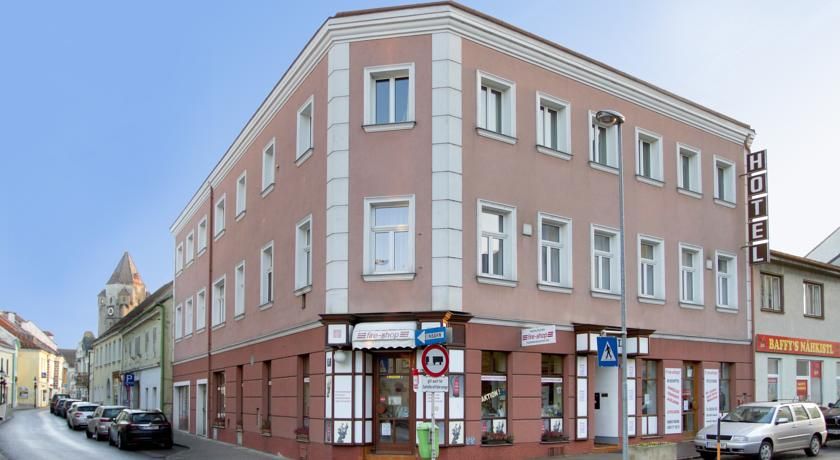 Háromemeletes szállodaépület egy utcasarkon, ahol autók és üzletek vannak.