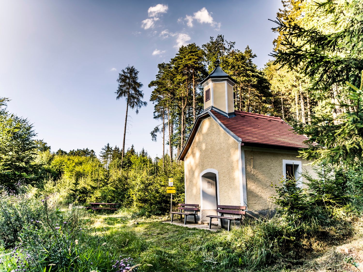 Kis kápolna az erdőben, előtte padokkal, fákkal és réttel körülvéve.